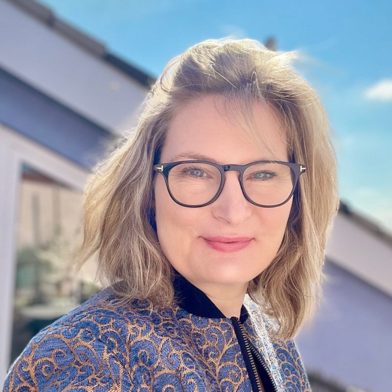 Frau mit blonden Haaren und Brille lächelt vor einer hellen, urbanen Kulisse. Digitale Produkte, Online Schulungen, Projektkoordination, VA