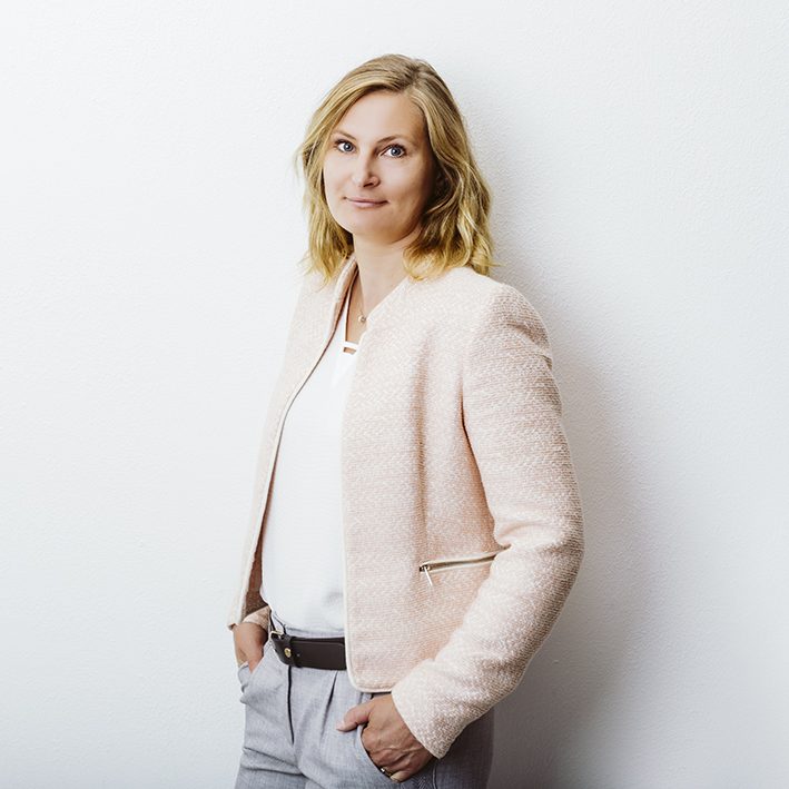 Eine blonde Frau in einem hellen Blazer steht entspannt vor einer weißen Wand.