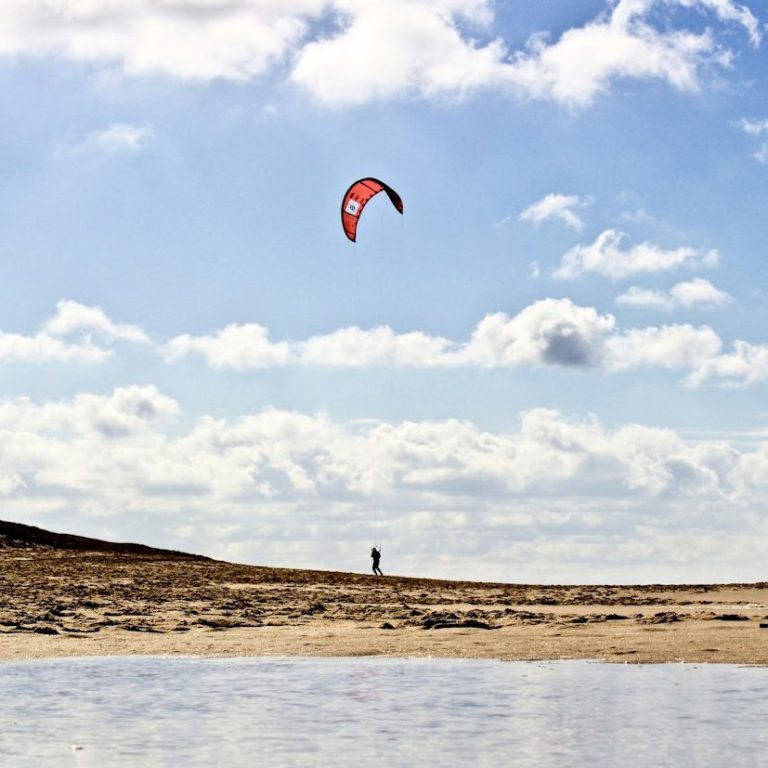 Kitesurfer vor einem weiten Strand unter einem hellem, wenig bewölkten Himmel.
