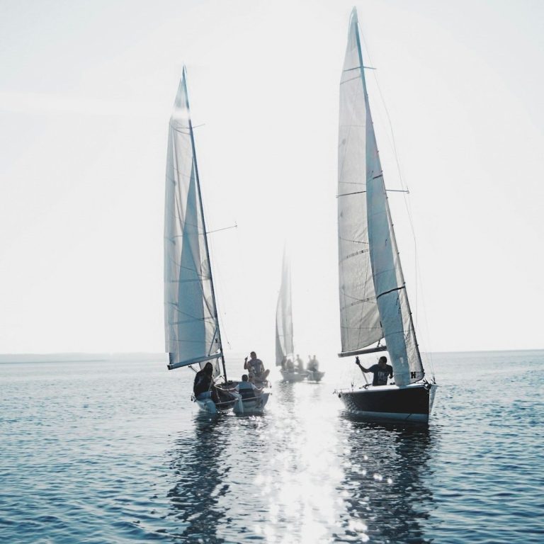 Segelboote bei Tageslicht fahren über ruhiges Wasser. Eins vorne, zwei dahinter mit einigen Personen, als Besatzung.